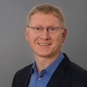 Porträt von Bernd Dinchel, lächelnder Mann mit Brille und dunklem Sakko vor grauem Hintergrund