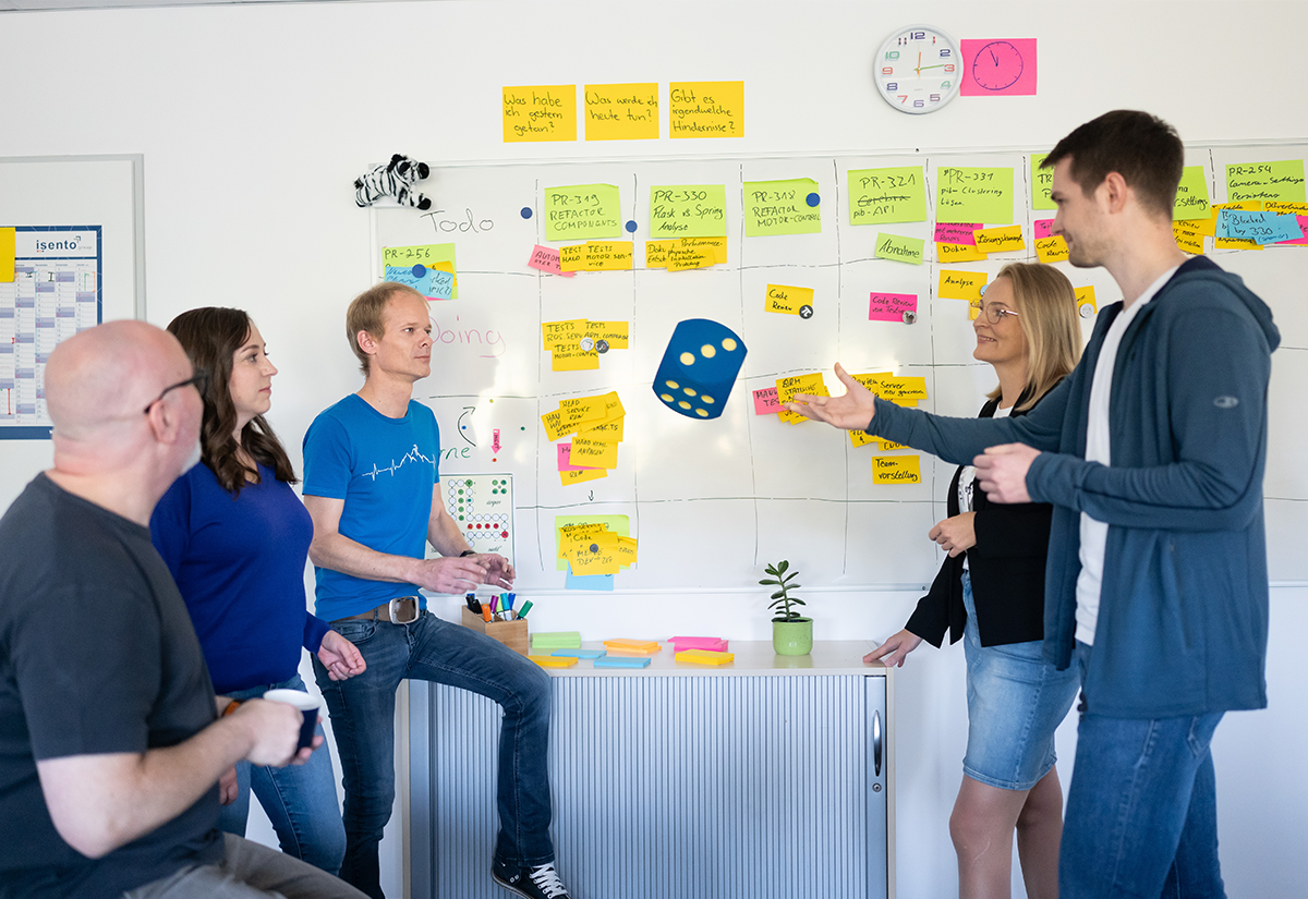 Team bespricht Aufgaben an einem Whiteboard, während ein großer Würfel geworfen wird