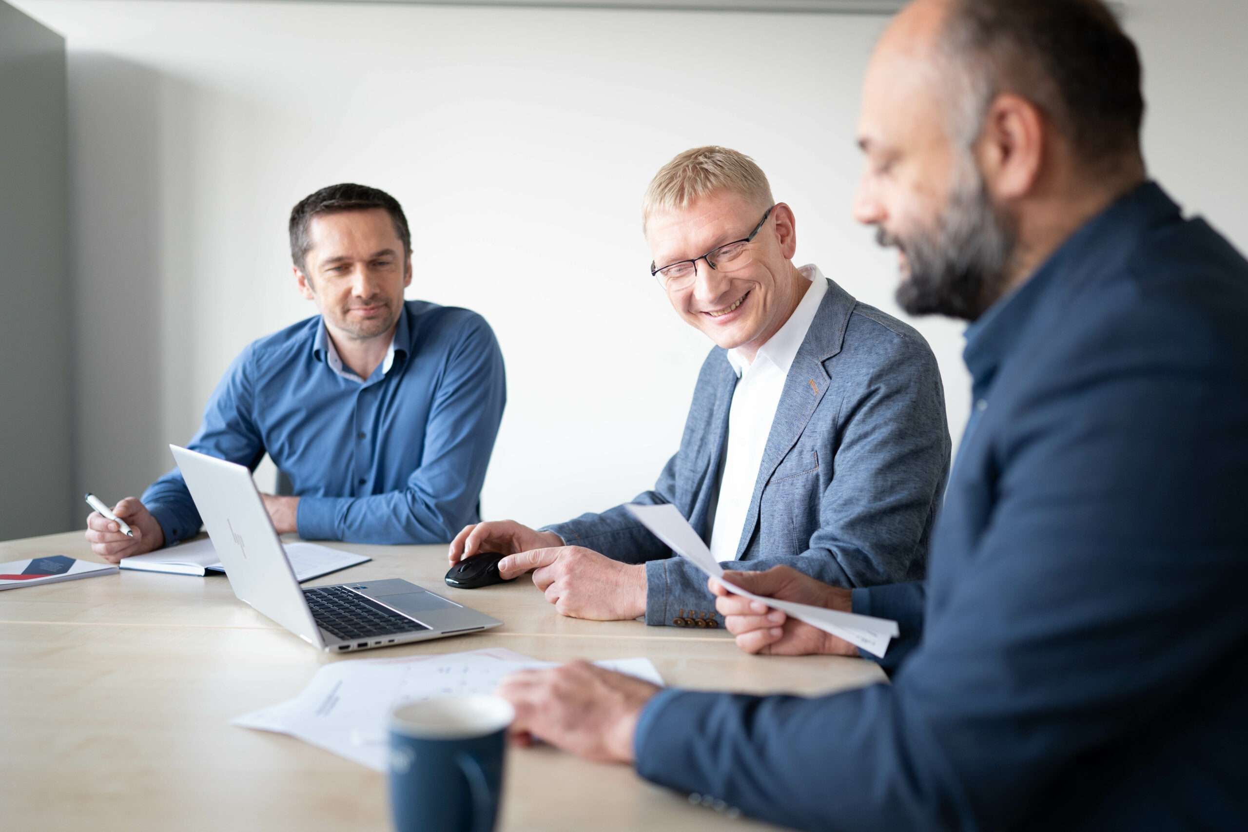 Drei Männer sitzen an einem Konferenztisch und arbeiten gemeinsam am Laptop und mit Unterlagen