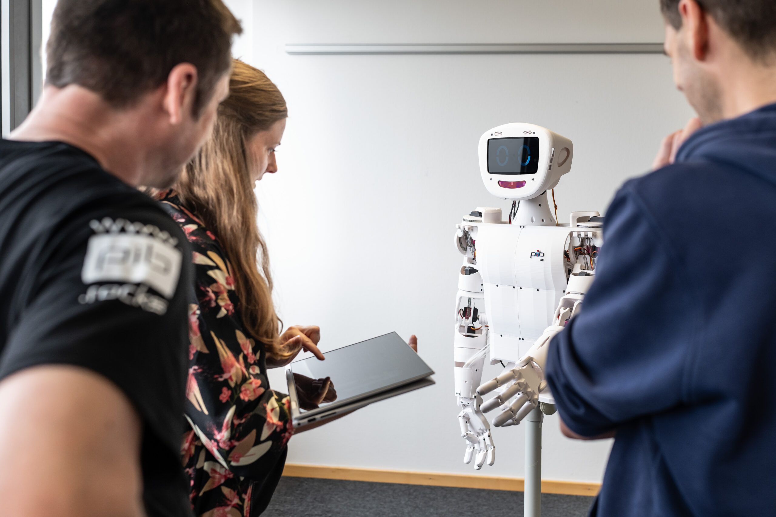 Drei Menschen stehen um einen humanoiden Roboter. Eine Frau hält ein Tablet in der Hand, um den Roboter zu steuern.